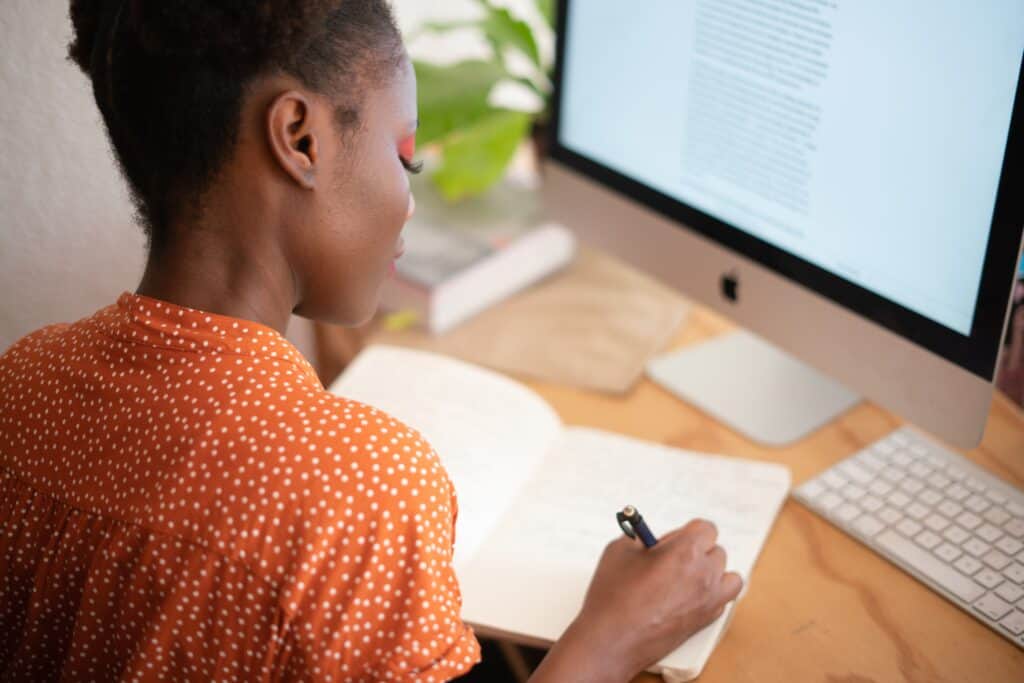 student writing in notebook with computer screen in front of her