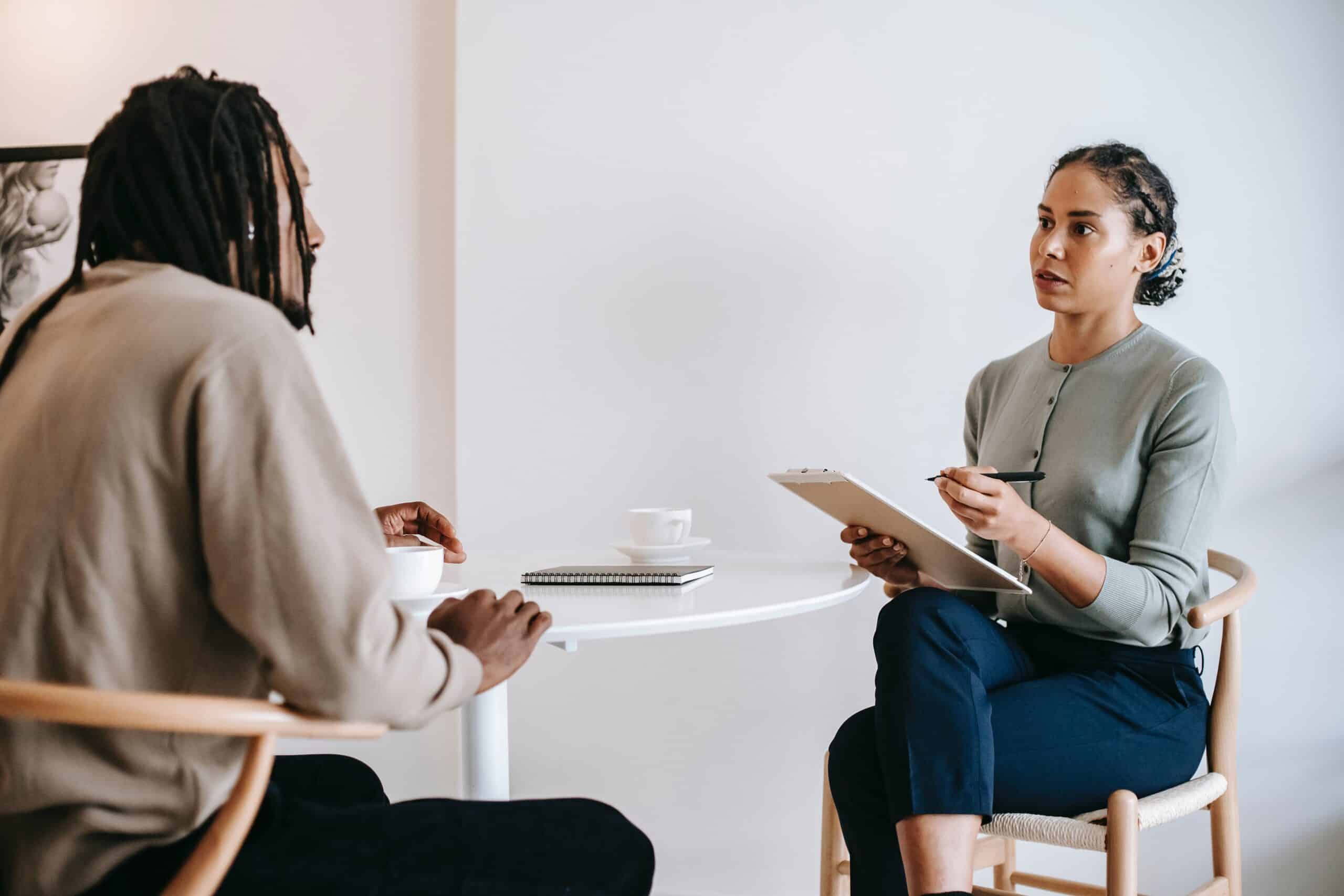 interviewer and interviewee sitting at table. Interviewer has clipboard in hand and is speaking.