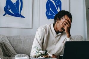 frustrated person with hand covering their facing sitting on couch in front of their laptop