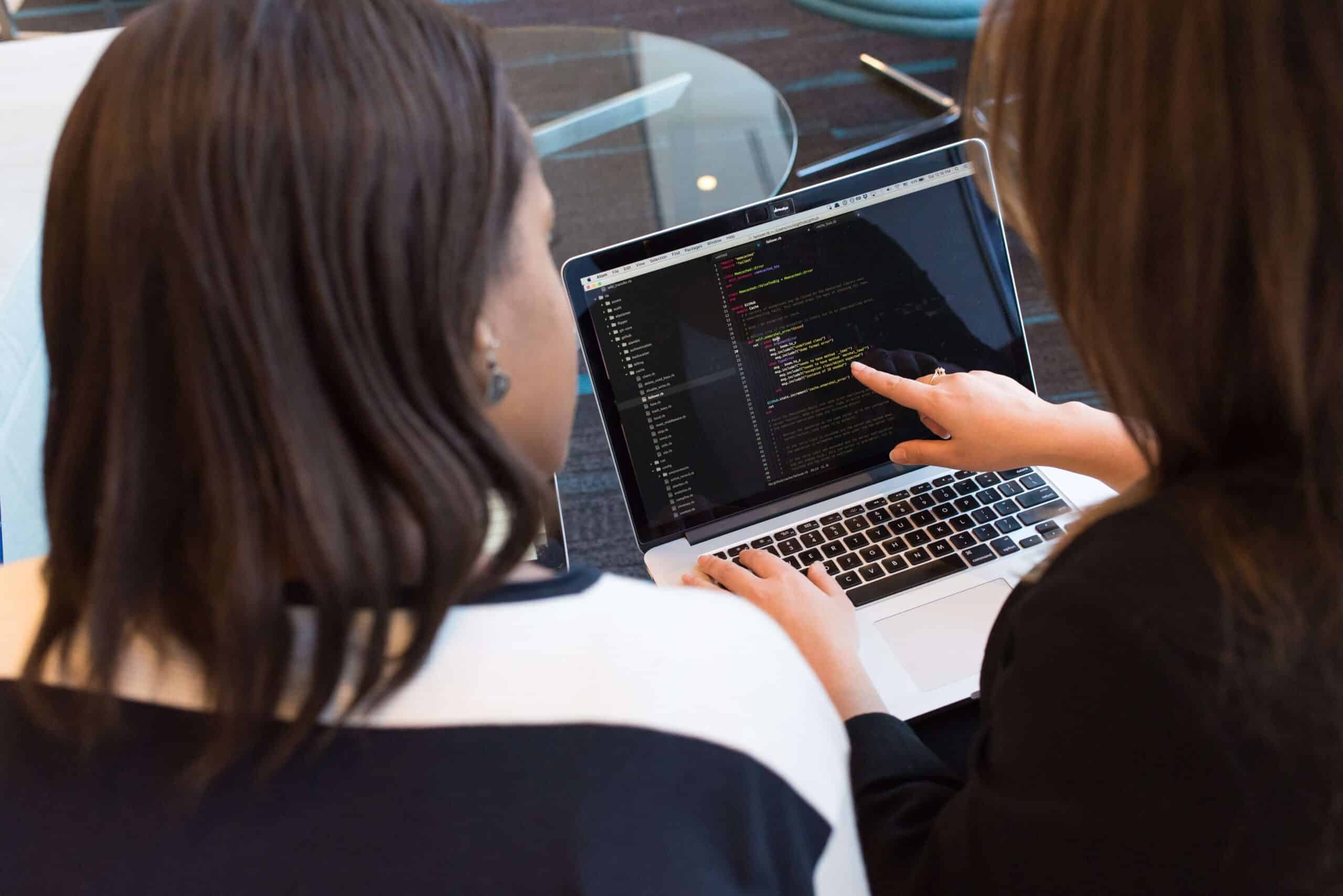 person pointing to code on laptop with other person looking on