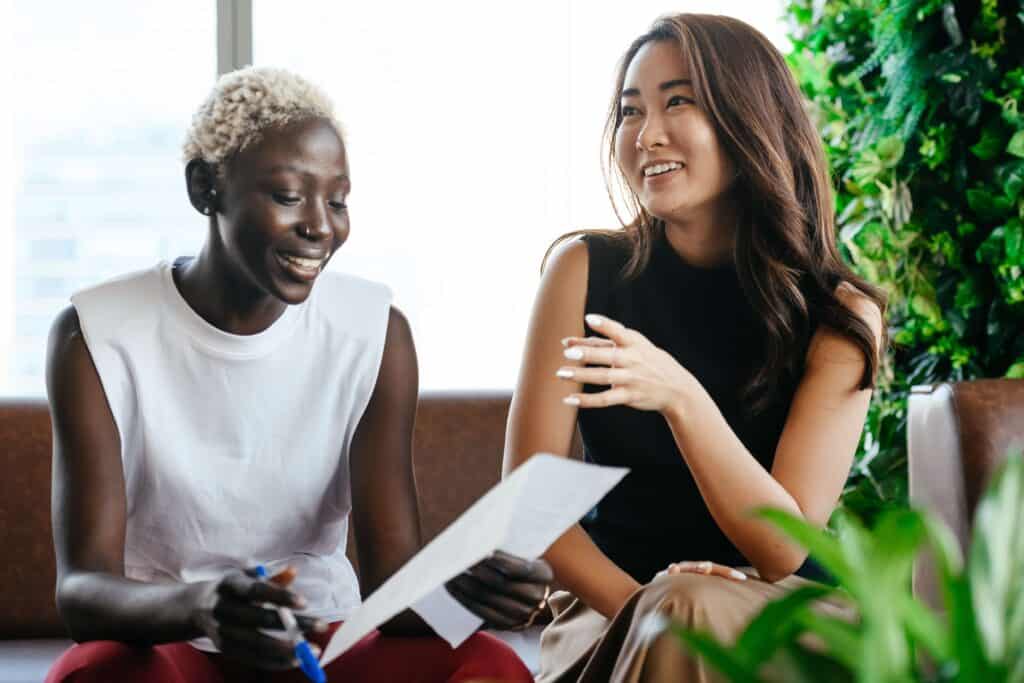 two women sitting on a bench, laughing and gesturing toward a document.